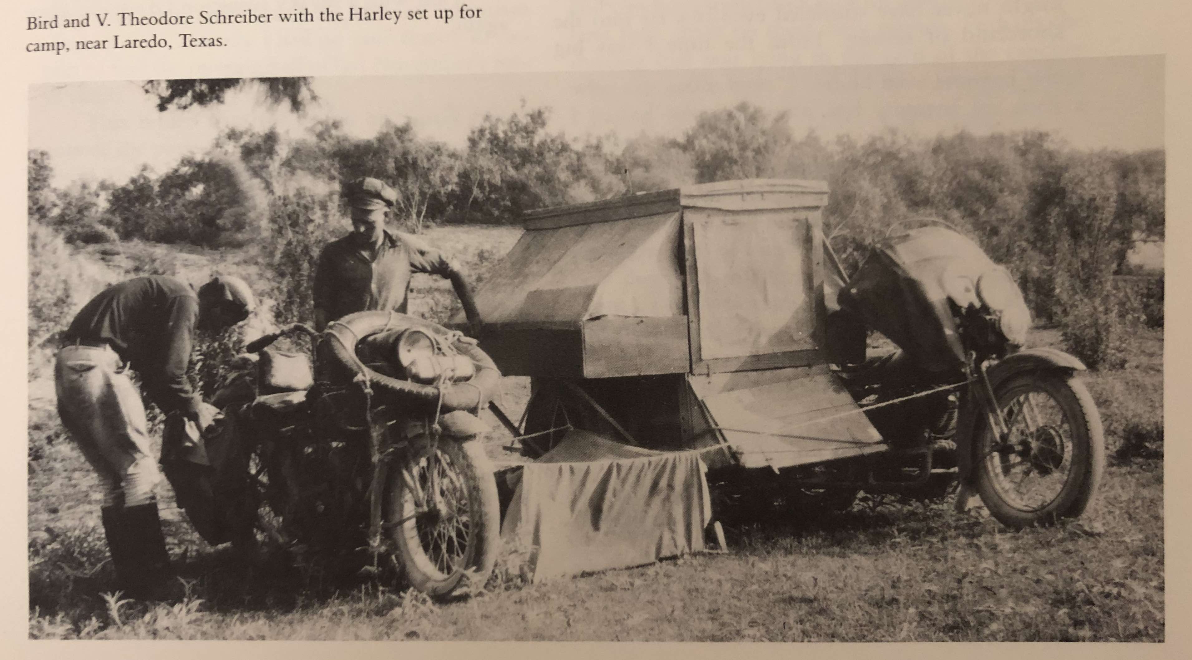 Black and white photo of two men. A canvas tent and a clothesline with a sheet hanging from it is stretched between two motorcycles which are absolutely loaded down with stuff, including several saddle bags and at least one spare tire. One of the men is fiddling with the motorcycle on the left. It's not clear to me which person is Bird.  