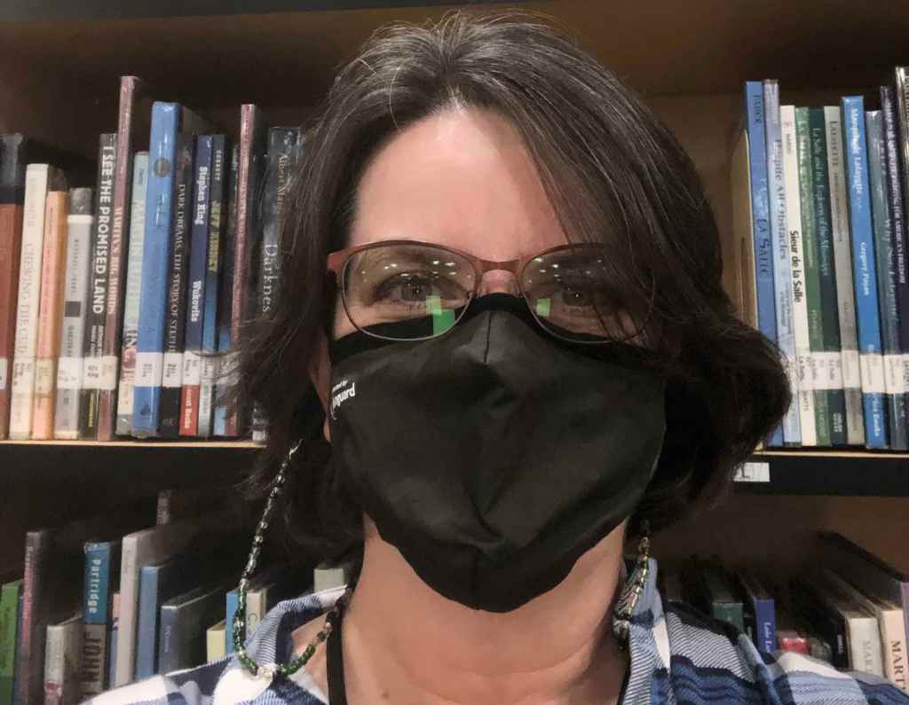 Me in a black mask, posing in front of a shelf of books, wearing a blue plaid shirt. 
